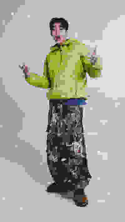 A person wearing a bright green puffy jacket and camouflage pants stands in a snowy environment, gesturing with their hands.