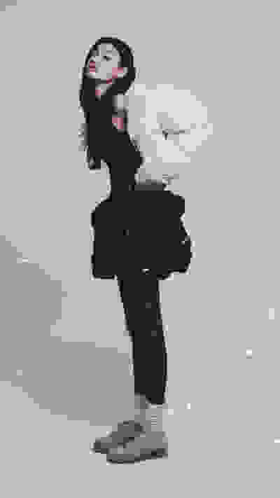 A woman in a black dress and white puffer jacket stands in a snowy outdoor setting, with snowflakes falling around her.