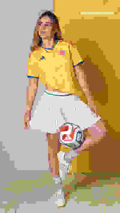 A young woman wearing a yellow soccer jersey and a white pleated skirt stands in front of a bright yellow background, holding a soccer ball.