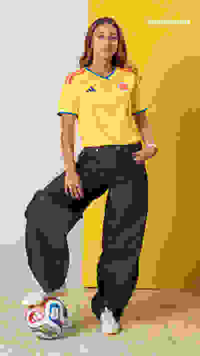 A young woman wearing a yellow soccer jersey and black pants stands in front of a yellow background, posing with a soccer ball at her feet.