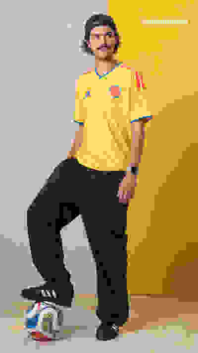 A man wearing a yellow soccer jersey and black pants is standing in front of a yellow background, with a soccer ball at his feet.