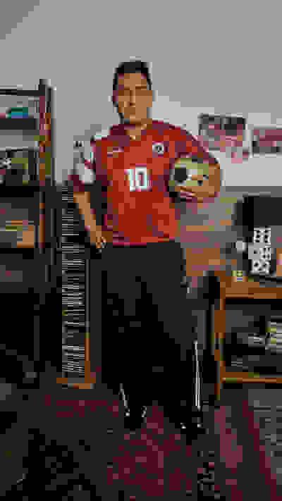 A young man wearing a red soccer jersey with the number 10 stands in a room filled with shelves of vinyl records and other memorabilia.
