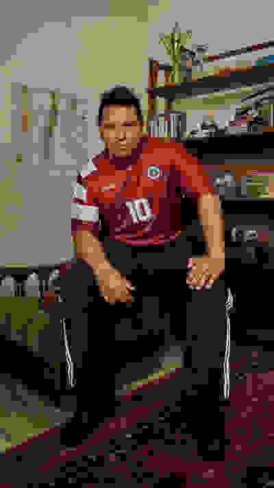 The image shows a man wearing a red sports jersey with the number 10 sitting on a couch in a room with bookshelves and a world map on the wall.