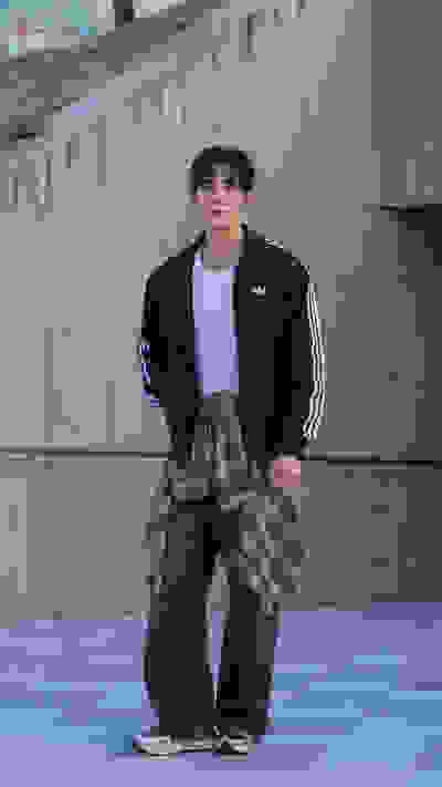 A young person wearing a black jacket and plaid pants stands in front of a concrete wall in an urban setting.