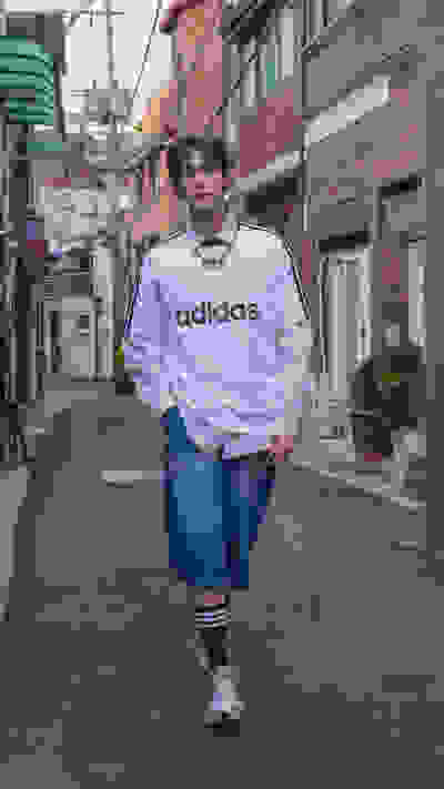 A person wearing an Adidas sweatshirt and jeans is standing on a street with brick buildings and storefronts in the background.