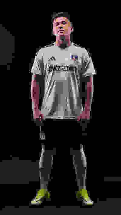A muscular male athlete wearing a white soccer jersey with the "Juga" logo, standing in a dark background and wearing bright green cleats.