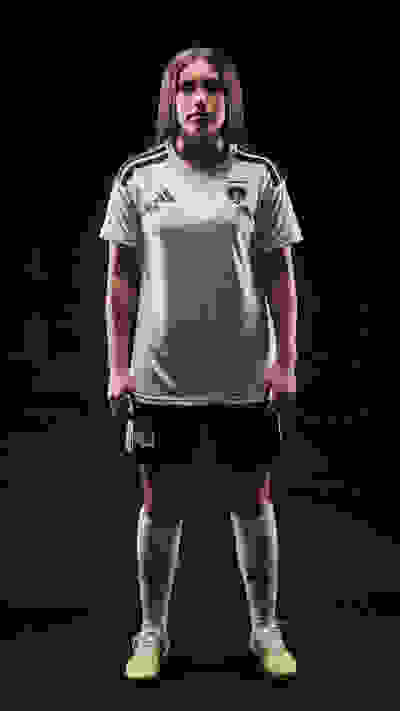A person wearing a gray soccer jersey and shorts, standing in a dark background.