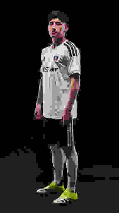 A young man wearing a white soccer jersey and black shorts stands against a dark background, his feet adorned with bright green cleats.