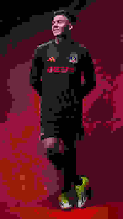 A young man wearing a black sports jersey with the team logo "Johor" stands against a deep red background, his bright yellow cleats adding a vibrant contrast to the scene.