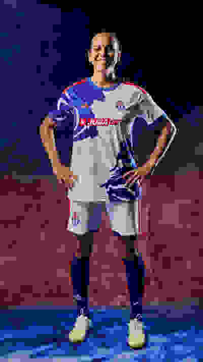 The image shows a smiling soccer player wearing a white and blue jersey, standing on a field with a red and blue background.