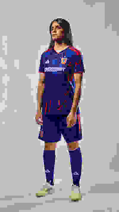 A young woman wearing a blue and red soccer jersey and shorts stands confidently against a plain background.