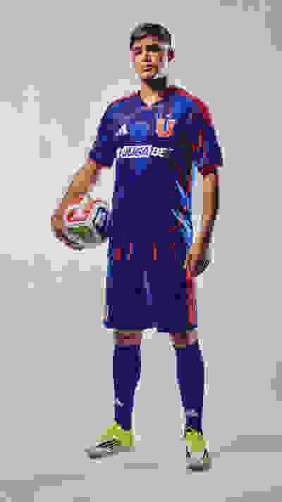 A man wearing a blue and red soccer jersey and shorts, holding a soccer ball, stands against a plain background.