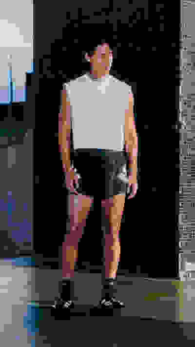 A muscular individual wearing a white sleeveless top and black shorts stands in front of a dark background, appearing to be in a fitness or training setting.