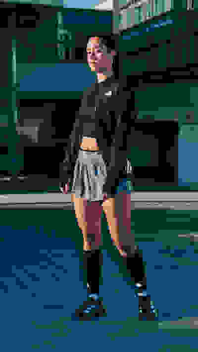 A young woman wearing a black crop top and gray shorts stands on a concrete surface, with a modern building in the background.