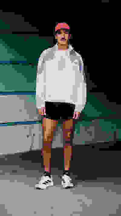 A person wearing a white sweatshirt, red cap, and sneakers stands in front of a green and blue wall.