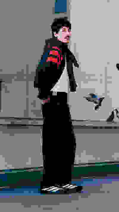 A person wearing a black and red striped jacket stands in front of a concrete wall, with pigeons visible in the background.