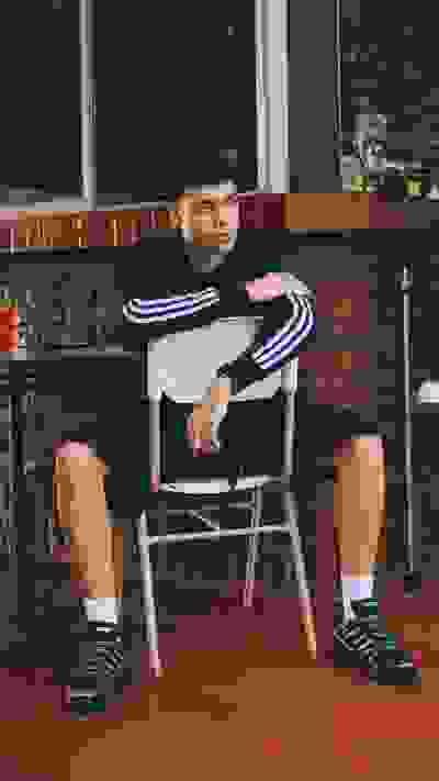 A young man in casual attire sits on a chair in what appears to be a restaurant or cafe setting, with a brick wall and other furnishings visible in the background.