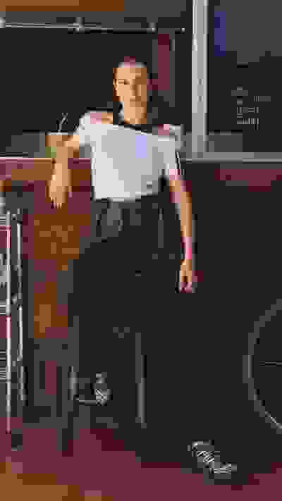 A young woman with dark hair sits on a stool in front of a brick wall, wearing a white top and dark pants, with a bicycle visible in the background.