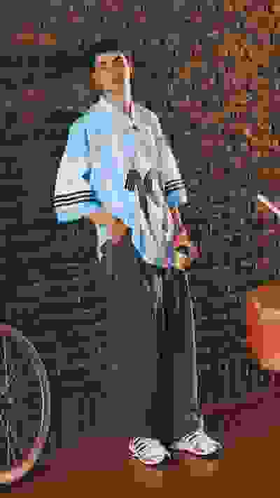A young man wearing a colorful striped shirt and loose-fitting pants stands in front of a brick wall, with a bicycle visible in the background.