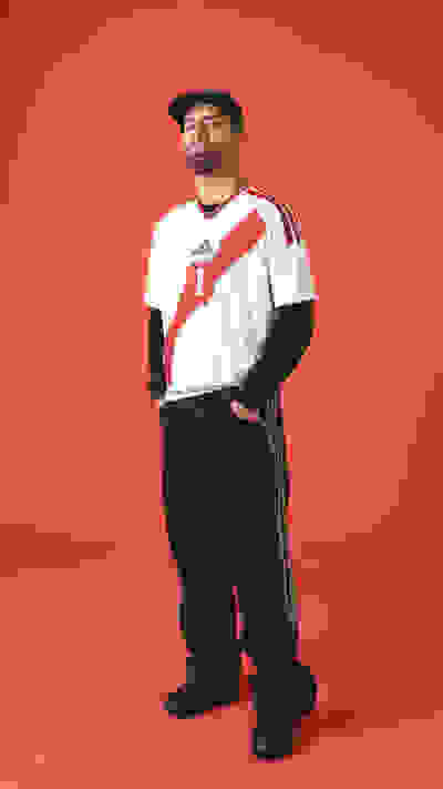 A person wearing a white sports jersey, black pants, and black shoes stands against a bright red background.