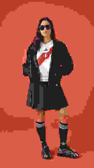 A young woman wearing a black jacket, white shirt, and black shorts stands against a bright red background, with sunglasses and athletic shoes completing her stylish outfit.