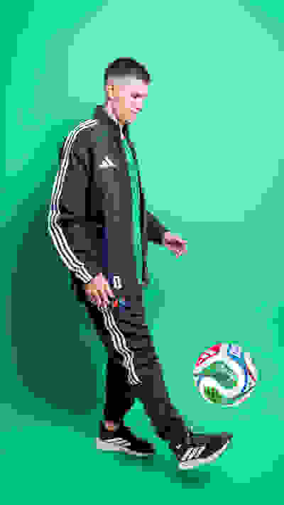 A young man in a sports jacket and pants is juggling a soccer ball against a vibrant green background.