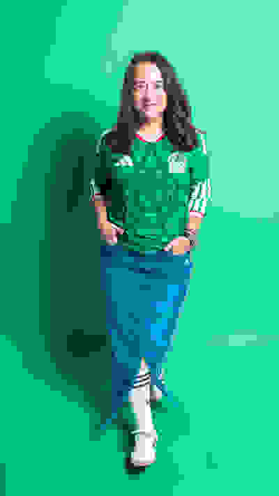 A young woman with long dark hair is standing in front of a vibrant green background, wearing a green jersey with a logo and smiling at the camera.
