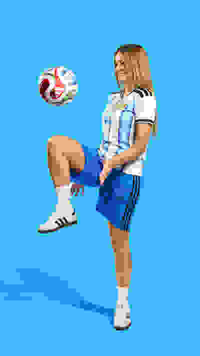 A young woman in a sports uniform is kicking a soccer ball against a bright blue background.