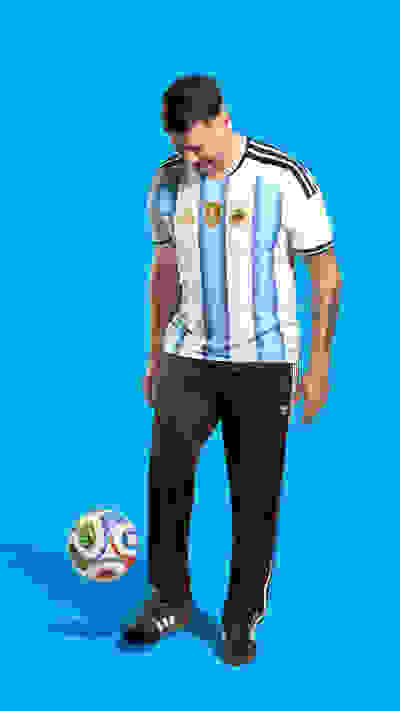 A muscular man wearing an Argentina national team jersey stands next to a soccer ball against a bright blue background.