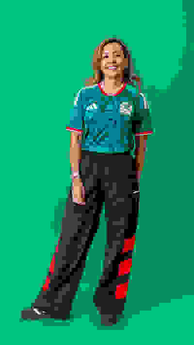 A smiling person wearing a green and white sports jersey and navy blue pants stands in front of a vibrant green background.