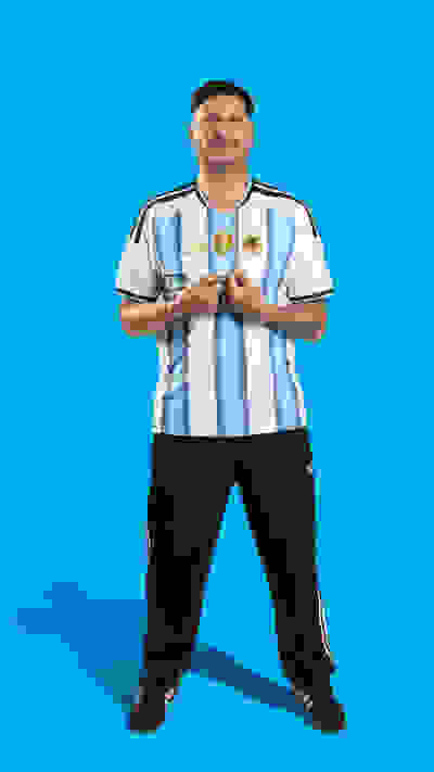 A smiling man wearing an Argentina national team jersey stands against a bright blue background.