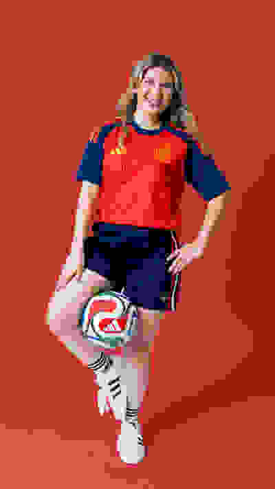 A smiling woman in a red and blue soccer jersey and shorts, holding a soccer ball, stands against a vibrant red background.