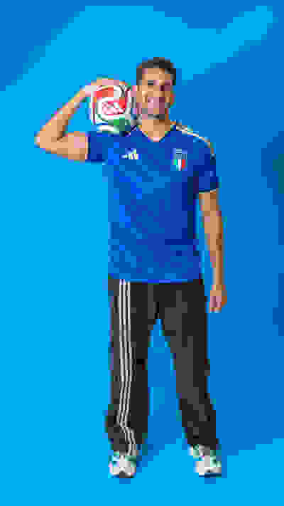 A smiling man wearing an Italian national team soccer jersey and holding a soccer ball stands in front of a vibrant blue background.