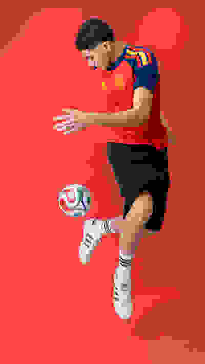 A young man in a red and blue soccer jersey is jumping and kicking a soccer ball against a bright red background.