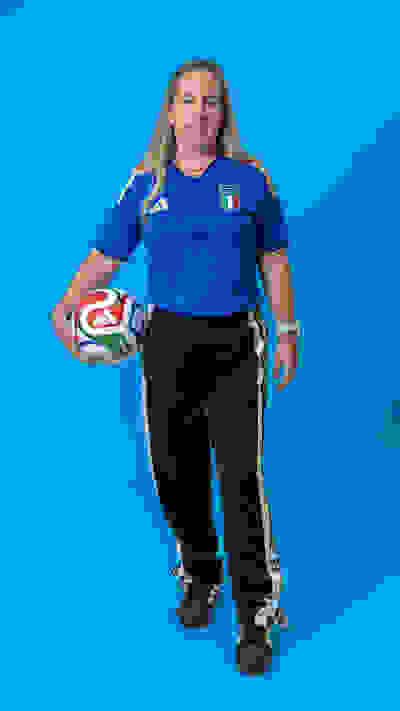 A young woman wearing a blue soccer jersey and holding a soccer ball stands in front of a bright blue background.