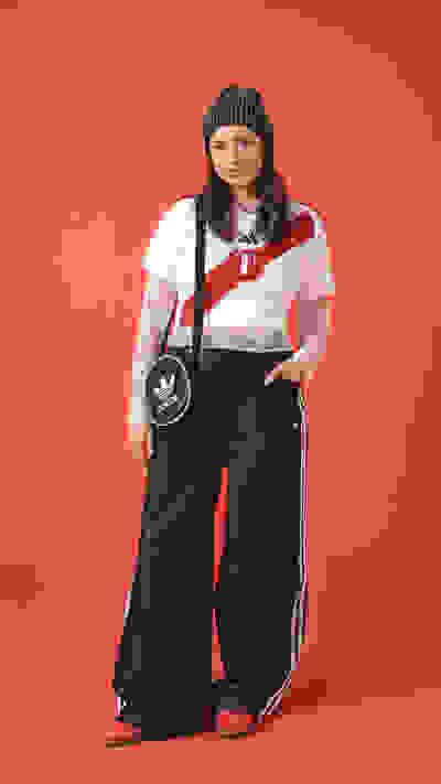 A young woman wearing a white shirt, black pants, and a striped hat stands against a bright red background.