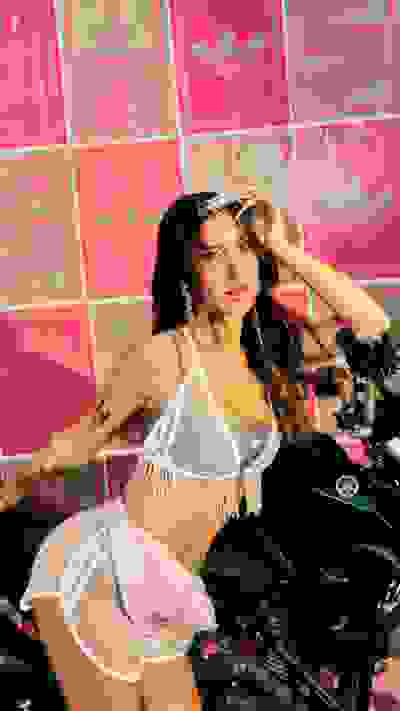 A woman in a white and pink outfit sits on a motorcycle in front of a vibrant pink and white background with the word "Carnaval" prominently displayed.