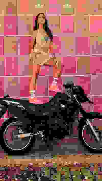 A woman in a revealing outfit poses on a motorcycle in front of a vibrant pink and white background adorned with Adidas logos.