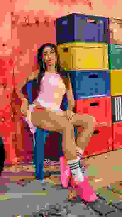 A young woman with long dark hair sits on a colorful chair in front of a vibrant red wall, surrounded by stacked plastic crates in various bright colors.