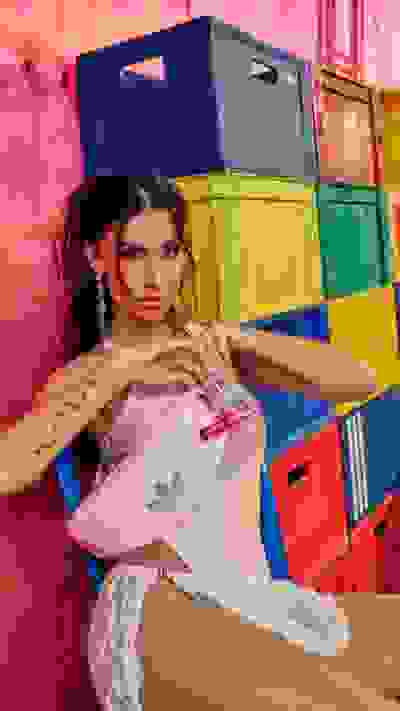 A young woman with curly hair and tattoos poses in front of a colorful backdrop of stacked crates.