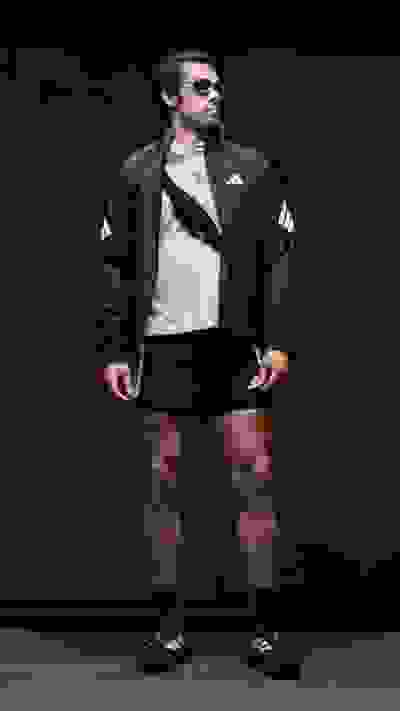 A man wearing a black jacket, white shirt, and black shorts stands in a dark setting, his muscular physique and confident posture commanding attention.