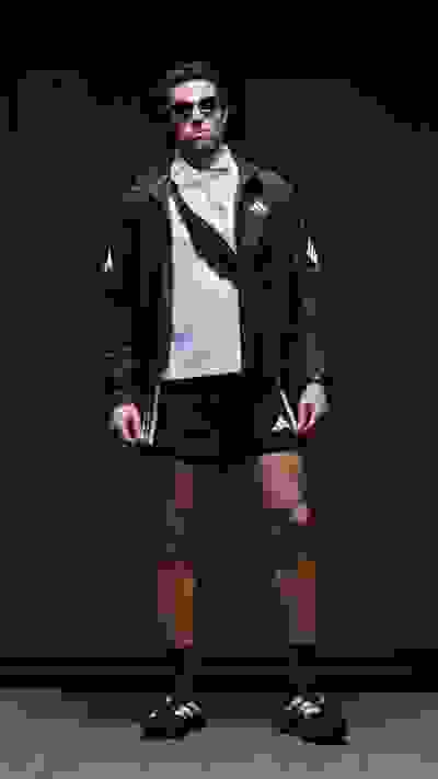 A person wearing a black jacket, white shirt, and black shorts stands in front of a dark background.