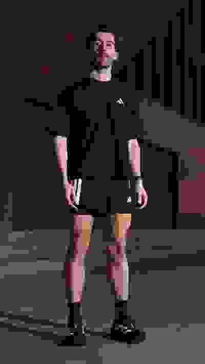 A muscular man wearing a black athletic outfit stands in a dimly lit indoor setting, with a plain background behind him.