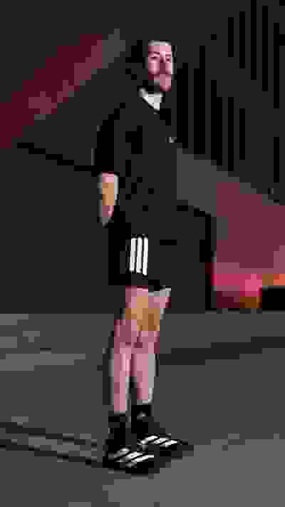 A person with a beard wearing a black shirt and shorts stands in a dimly lit area with a patterned wall in the background.