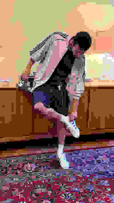 A person with curly hair is sitting on a wooden cabinet, wearing a light-colored jacket, shorts, and white sneakers, against a background of a patterned rug and a lamp on the cabinet.