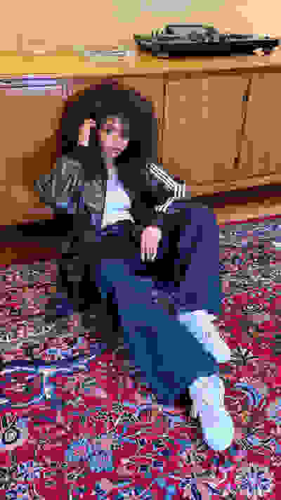 A young person with curly hair sits on a colorful patterned rug, surrounded by a wooden cabinet and audio equipment in the background.