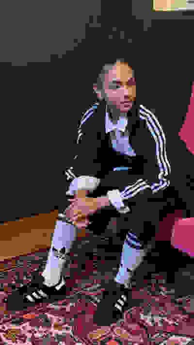 A young person with curly hair wearing a school uniform sits on a patterned rug in a room with dark walls.