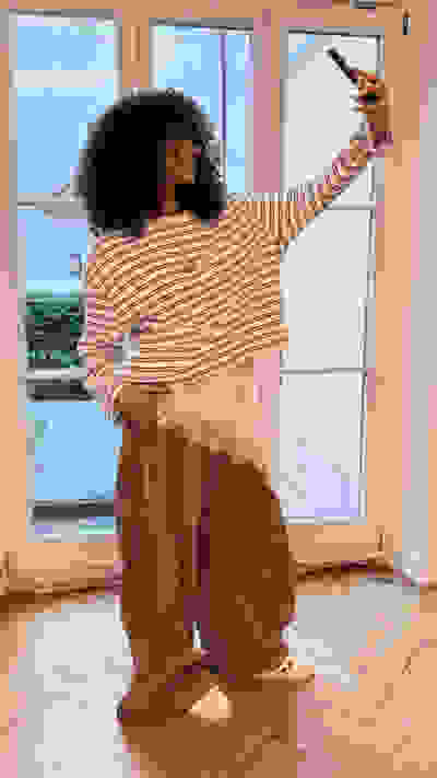 A person with curly hair wearing a striped shirt and brown pants stands in a room with large windows overlooking a scenic outdoor view.