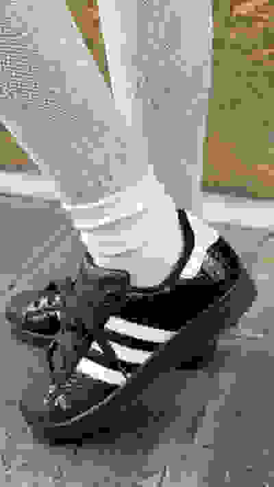 The image shows a pair of black and white sneakers with a lace-up design, worn with white lace-patterned stockings against a concrete background.