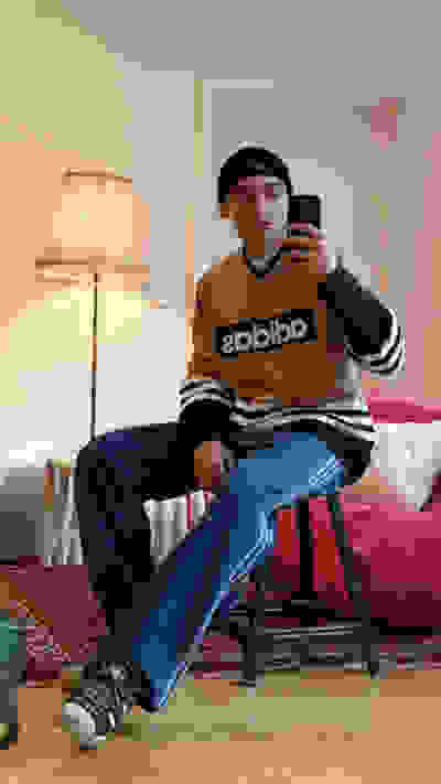 A person wearing an Adidas sweatshirt is sitting on a chair in a room with a lamp and a patterned rug in the background.
